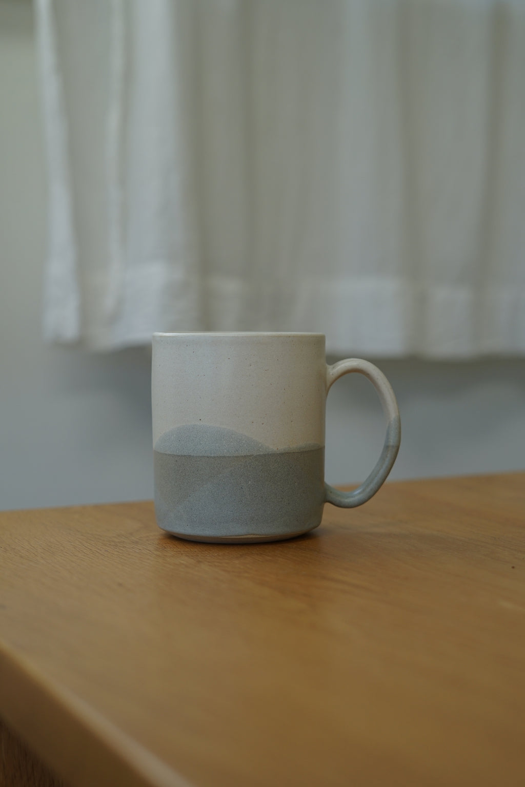 A ceramic mug with a two-tone design, featuring a white upper half and a textured light gray lower half, sitting on a wooden table with a soft-focus white curtain background.