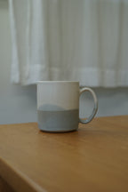 A ceramic mug with a two-tone design, featuring a white upper half and a textured light gray lower half, sitting on a wooden table with a soft-focus white curtain background.
