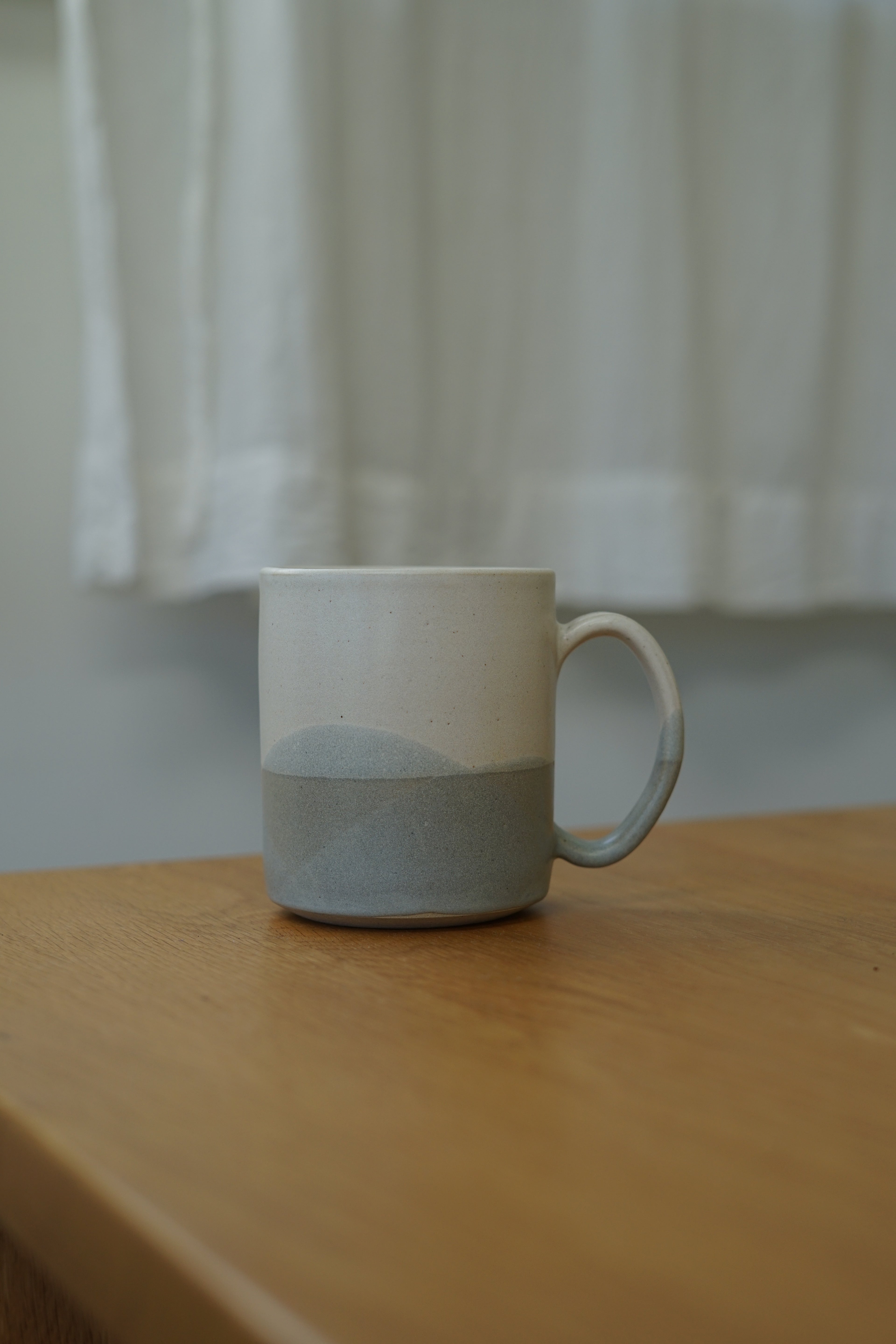 A ceramic mug with a two-tone design, featuring a white upper half and a textured light gray lower half, sitting on a wooden table with a soft-focus white curtain background.
