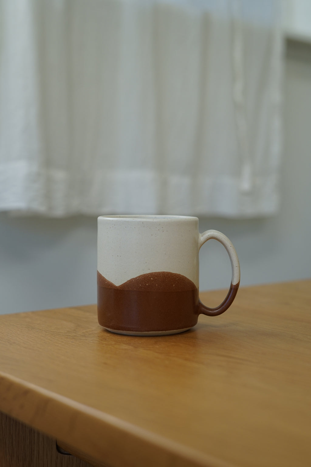 A ceramic mug with a two-tone design, featuring a white upper half and a brown lower half, sitting on a wooden table with a softly blurred white curtain background.
