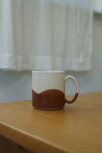 A ceramic mug with a two-tone design, featuring a white upper half and a brown lower half, sitting on a wooden table with a softly blurred white curtain background.